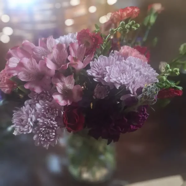 Lavender Lush bouquet featuring pink alstroemerias, lavender chrysanthemums, red roses, and mixed blossoms in a glass vase with warm bokeh background