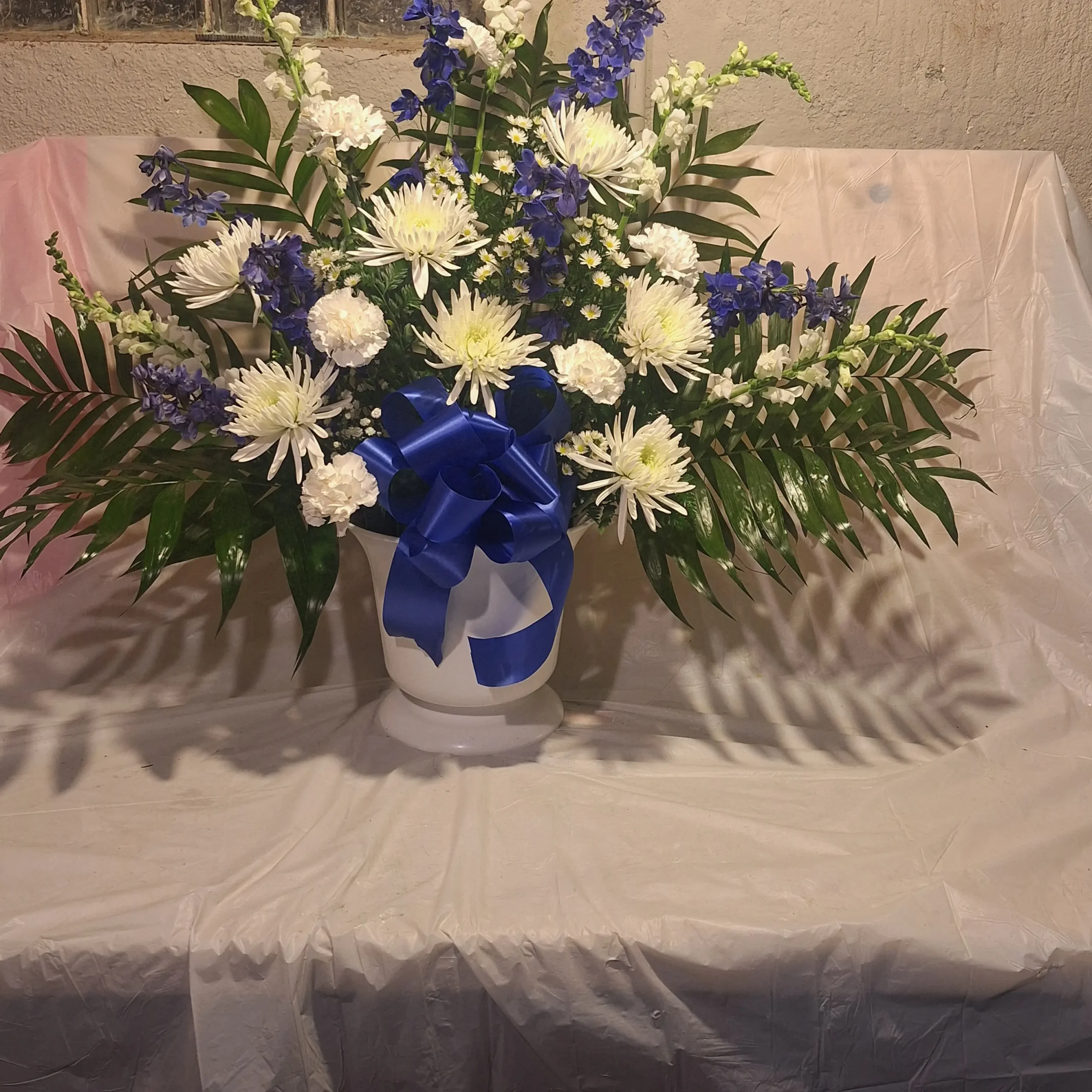 Large blue and white floral pedestal arrangement with white chrysanthemums, white carnations, blue delphiniums, and lush greenery in a white vase tied with a blue ribbon.