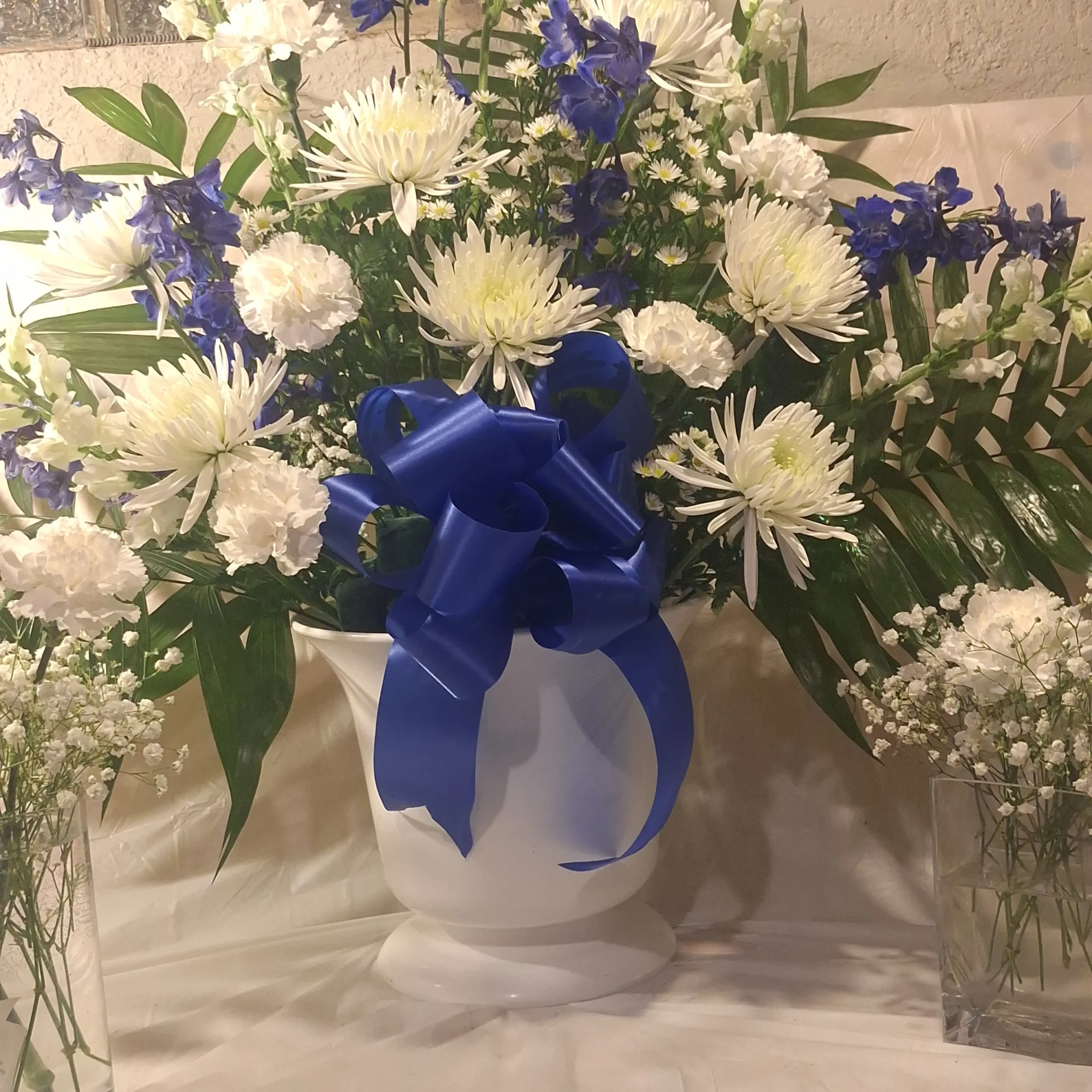 Large blue and white floral pedestal arrangement with white chrysanthemums, white carnations, blue delphiniums, and lush greenery in a white vase tied with a blue ribbon.
