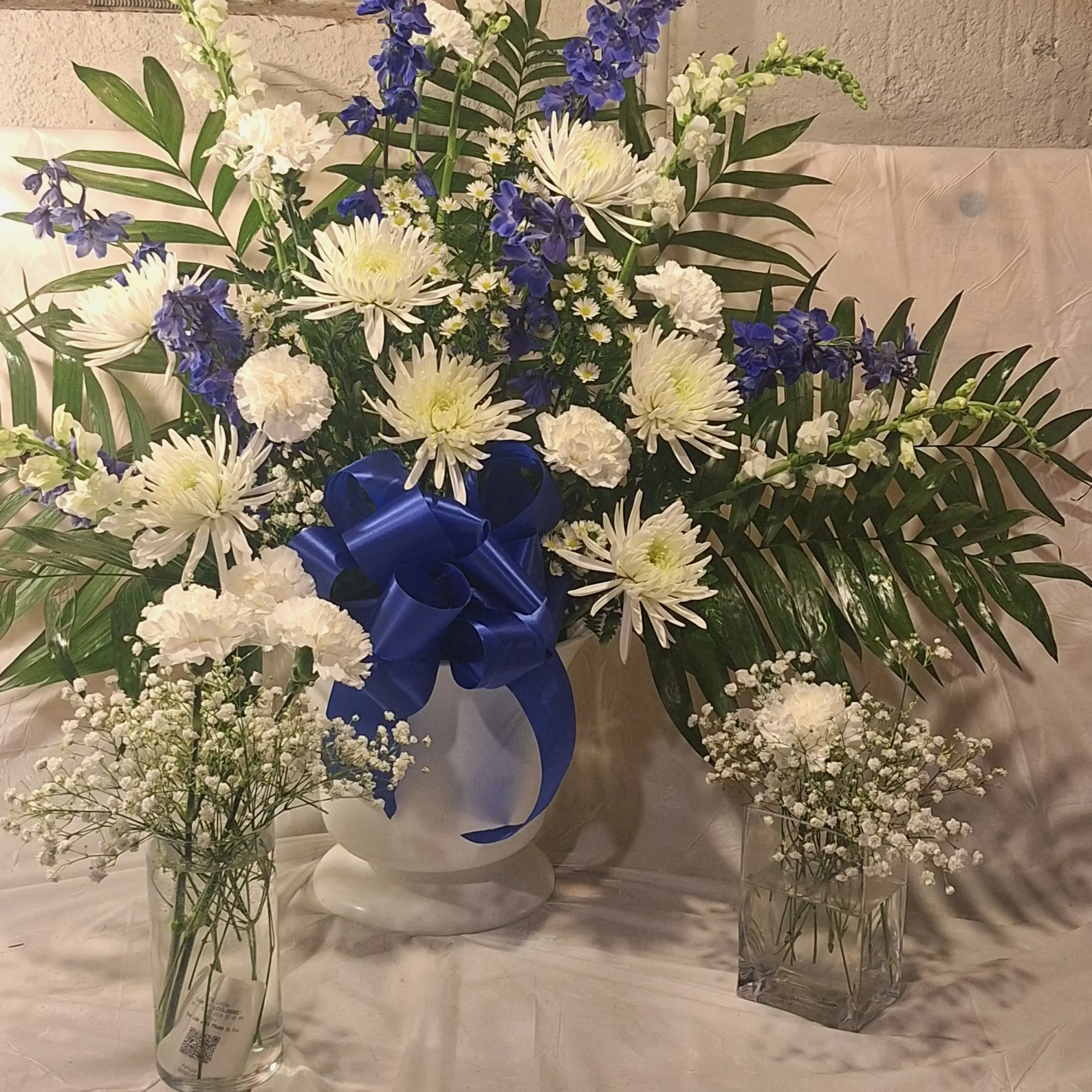 Large blue and white floral pedestal arrangement with white chrysanthemums, white carnations, blue delphiniums, and lush greenery in a white vase tied with a blue ribbon.