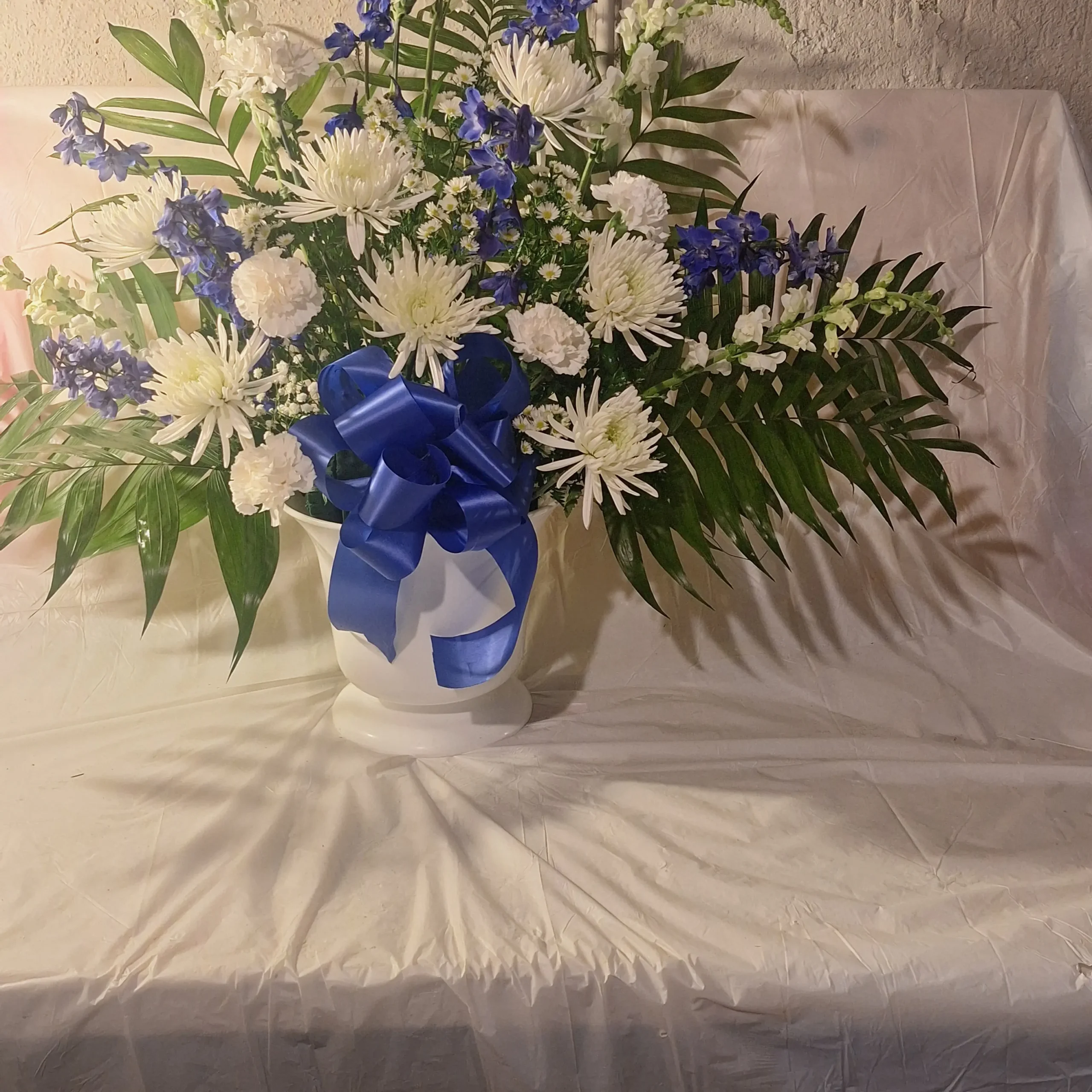 Large blue and white floral pedestal arrangement with white chrysanthemums, white carnations, blue delphiniums, and lush greenery in a white vase tied with a blue ribbon.