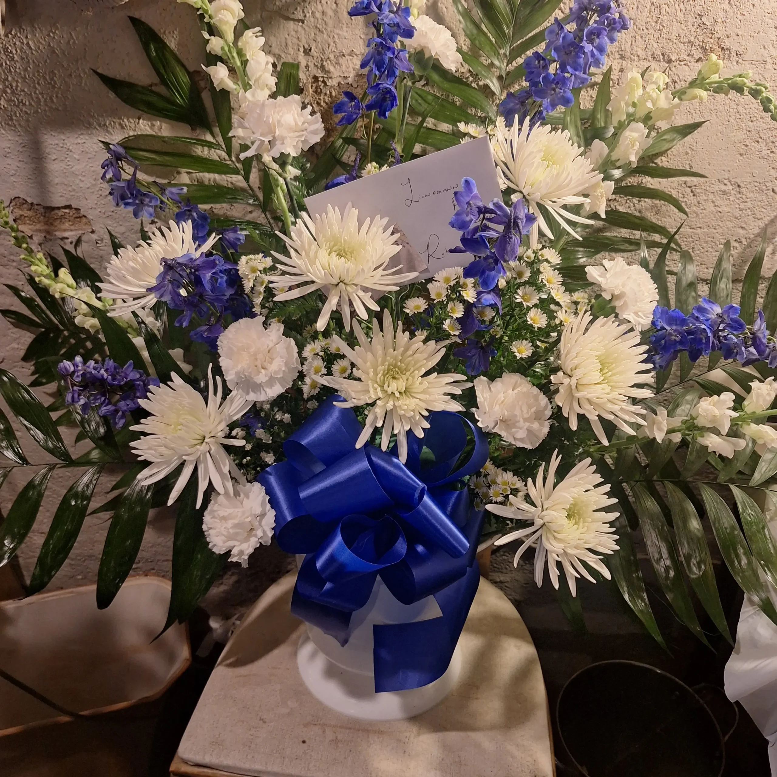 Large blue and white floral pedestal arrangement with white chrysanthemums, white carnations, blue delphiniums, and lush greenery in a white vase tied with a blue ribbon.