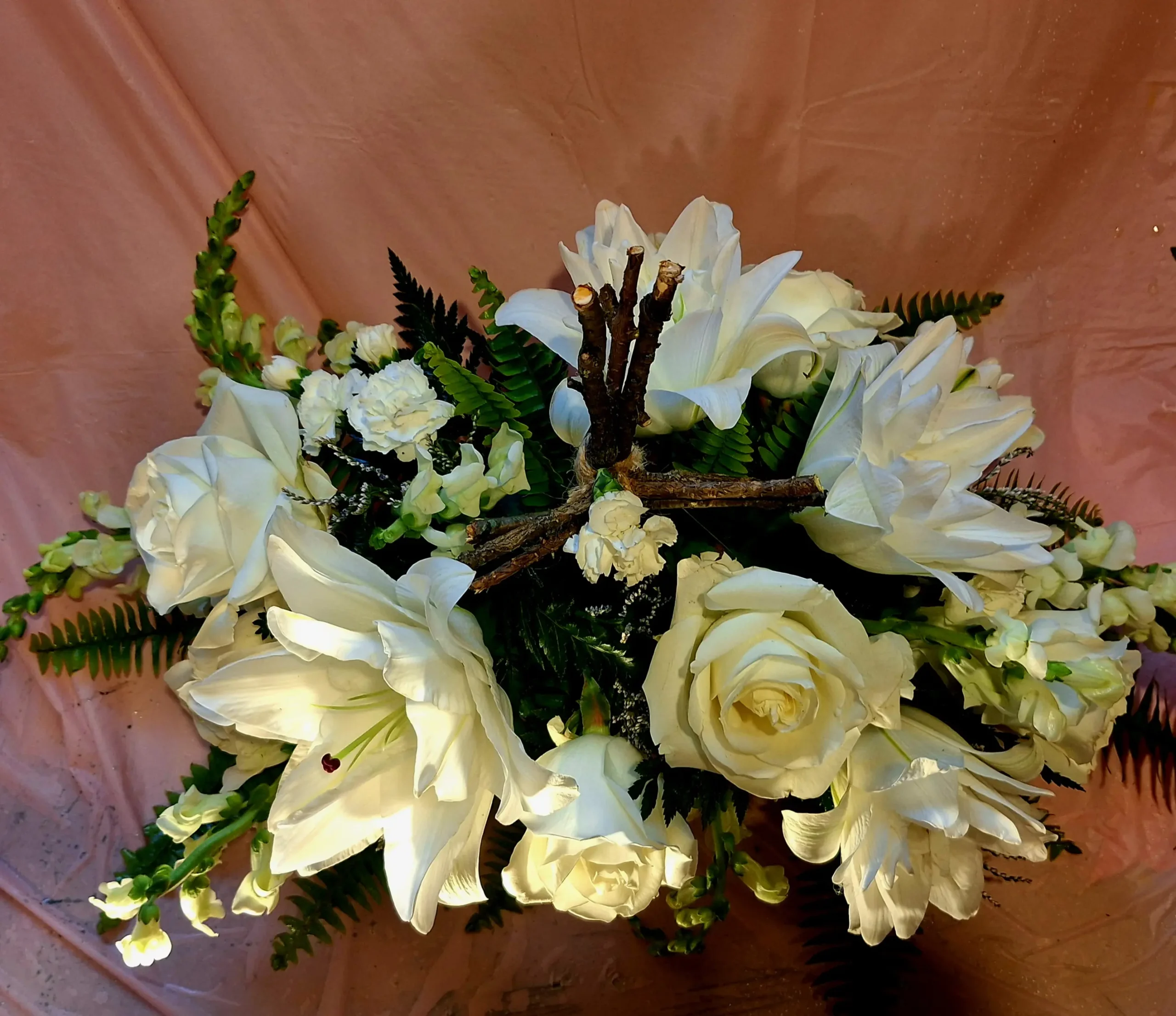 White roses, lilies, and carnations arranged with green ferns surrounding a rustic twig cross on a soft pink background.