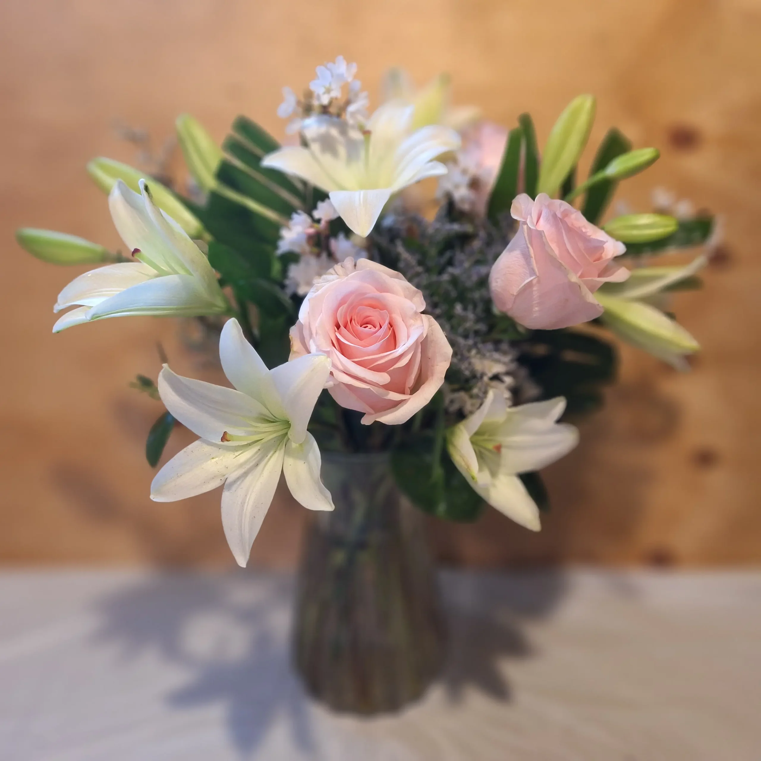 Blush roses, white lilies, and delicate white filler arranged in a clear glass vase with fresh greenery against a wooden backdrop.