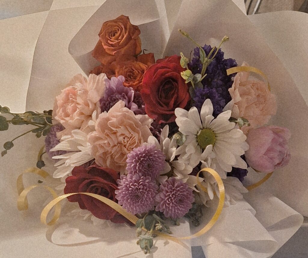 Bright multi‑color Mother’s Day floral arrangement in a short cubic vase featuring carnations, red roses, lavender mums, dark purple stock, tulips, spray roses, a daisy, and eucalyptus.