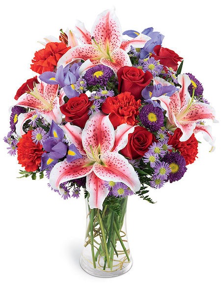 Clear glass vase with pink lilies, red roses, carnations, purple irises, and daisy‑like blooms arranged fresh with green foliage for Cincinnati flower delivery.