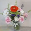 Blush and Bloom Elegance bouquet with pink roses, white daisies, orange gerbera daisy, and seasonal accents in a glass vase.