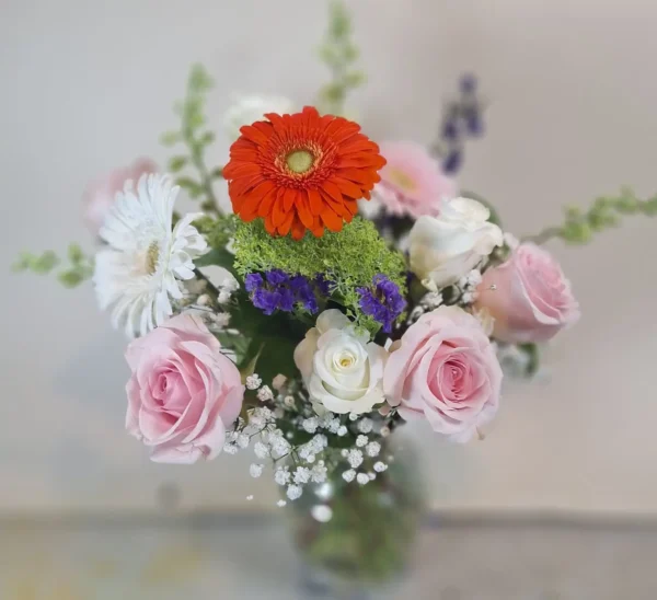 Blush and Bloom Elegance bouquet with pink roses, white daisies, orange gerbera daisy, and seasonal accents in a glass vase.