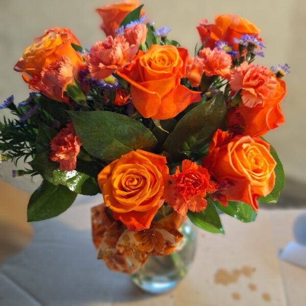 : Bright orange rose bouquet with pink carnations, purple fillers, and green leaves in a glass vase tied with an orange ribbon.