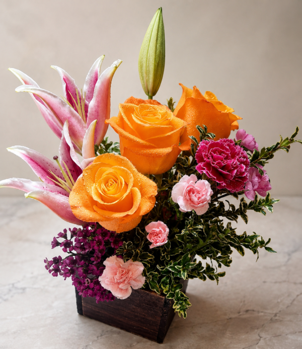 Amber Bloom Sonata floral arrangement with orange roses, pink lilies, magenta carnations, purple fillers, and variegated greenery in a dark rectangular container.