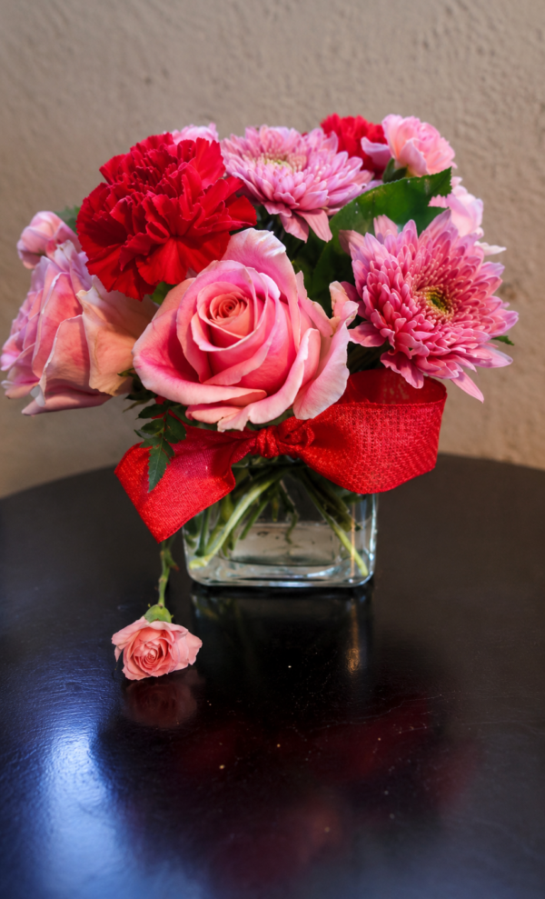 Petals of Promise Pink roses, red carnations, and pink chrysanthemums in a clear glass vase with red ribbon bow—fresh bouquet for Cincinnati flower delivery.
