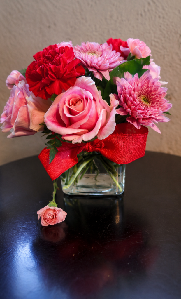 Pink roses, red carnations, and pink chrysanthemums in a clear glass vase with red ribbon bow—fresh bouquet for Cincinnati flower delivery.