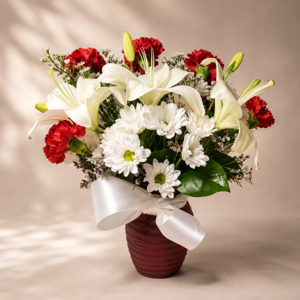 Elegant floral arrangement with white lilies, daisies, and red carnations in a red vase with satin ribbon