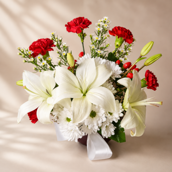 Elegant floral arrangement with white lilies, daisies, and red carnations in a red vase with satin ribbon