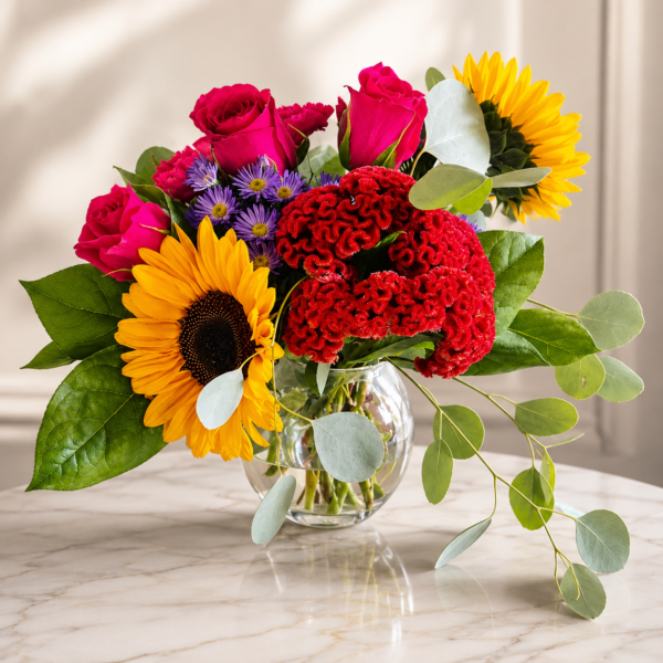 Bright bouquet of sunflowers, roses, cockscomb, asters, and eucalyptus in a clear glass vase on marble surface