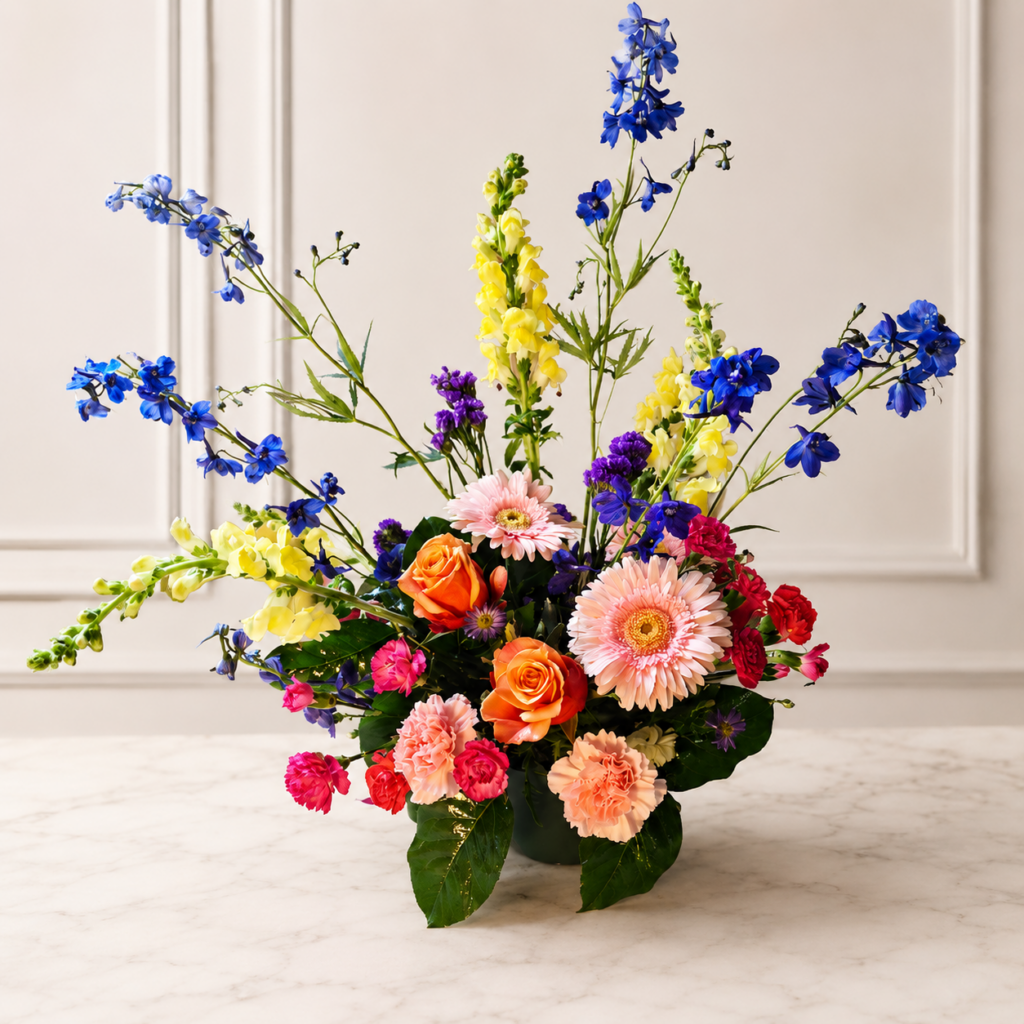 Cincinnati Colorburst bouquet with orange roses, pink gerbera daisies, peach carnations, bright pink mini carnations, yellow snapdragons, blue delphiniums, and purple accents, delivered fresh across Cincinnati and Northern Kentucky by allthepeoplesflowers.com
