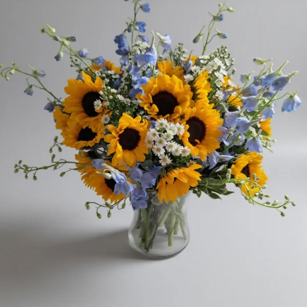 Morning Walk bouquet with sunflowers, blue delphinium, and white daisies in a clear glass vase.