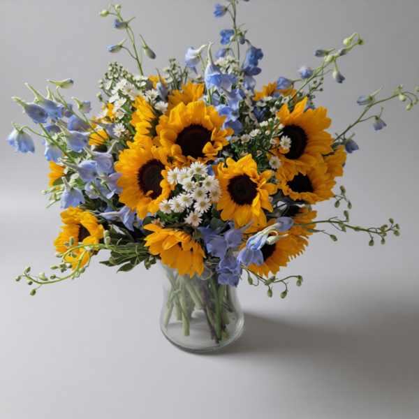 Morning Walk bouquet with sunflowers, blue delphinium, and white daisies in a clear glass vase.