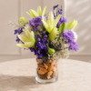 Seaside Breeze vase with light green lilies, purple chrysanthemums, deep purple delphiniums, baby’s breath, and decorative seed pods in a clear glass vase.