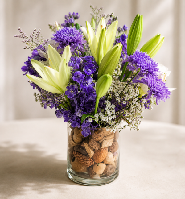 Seaside Breeze vase with light green lilies, purple chrysanthemums, deep purple delphiniums, baby’s breath, and decorative seed pods in a clear glass vase.