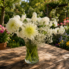 Elegant bouquet of white chrysanthemums, carnations, and baby’s breath in a clear glass vase on a wooden table with a garden background.