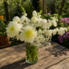 Elegant bouquet of white chrysanthemums, carnations, and baby’s breath in a clear glass vase on a wooden table with a garden background.