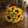 Fresh bouquet of sunflowers, white daisies, and greenery wrapped in gray and gold paper, displayed on a rustic wooden surface with warm indoor light.