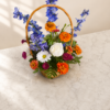 Wildflower Season Zinnia Basket with bright zinnias, delphiniums, roses, daisies, and greenery arranged in a woven basket on marble surface.