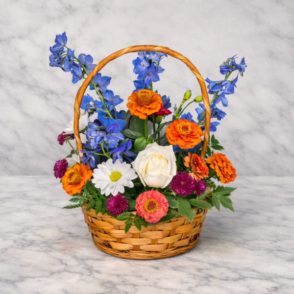 Wildflower Season Zinnia Basket with bright zinnias, delphiniums, roses, daisies, and greenery arranged in a woven basket on marble surface.