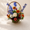 Wildflower Season Zinnia Basket with bright zinnias, delphiniums, roses, daisies, and greenery arranged in a woven basket on marble surface.