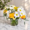 A Lovely Day bouquet with yellow roses, white daisies, filler blooms, and green foliage in a clear vase on a marble surface.