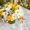 A Lovely Day bouquet with yellow roses, white daisies, filler blooms, and green foliage in a clear vase on a marble surface.