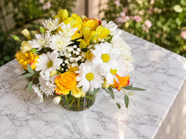 A Lovely Day bouquet with yellow roses, white daisies, filler blooms, and green foliage in a clear vase on a marble surface.