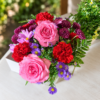 Amariah’s American Paradise bouquet with pink roses, red carnations, purple chrysanthemums, lavender daisies, and blue asters in a clear vase on a wooden table outdoors.