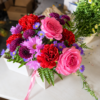 Amariah’s American Paradise bouquet with pink roses, red carnations, purple chrysanthemums, lavender daisies, and blue asters in a clear vase on a wooden table outdoors.