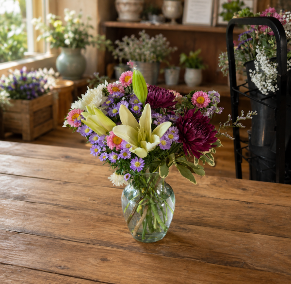 Autumn Sunset Designers Choice bouquet with chrysanthemums, lilies, carnations, and daisies in warm sunset tones, handcrafted in a clear glass vase.