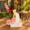 Coffee and Flowers To Go arrangement with pink carnations, red rose, purple asters, and tall blossoms in a white container with yellow ribbon, placed on a café table.