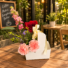 Coffee and Flowers To Go arrangement with pink carnations, red rose, purple asters, and tall blossoms in a white container with yellow ribbon, placed on a café table.