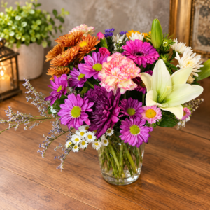 Fall Colors Designers Choice bouquet with lilies, chrysanthemums, carnations, and daisies in warm autumn tones, arranged in a clear glass vase.