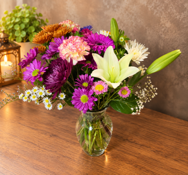 Fall Colors Designers Choice bouquet with lilies, chrysanthemums, carnations, and daisies in warm autumn tones, arranged in a clear glass vase.