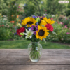 Sunflowers and Lily Celebration bouquet with bright sunflowers, white lilies, red carnations, and purple blooms in a clear vase on a wooden table outdoors.
