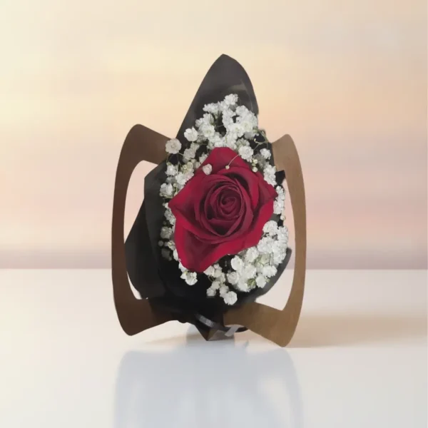 A single red rose wrapped in black and brown paper, surrounded by white baby’s breath, resting on a white surface with a softly blurred warm background.