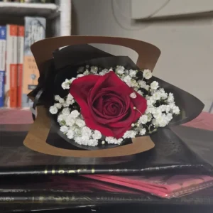 A single red rose wrapped in black and brown paper, surrounded by white baby’s breath, resting on a white surface with a softly blurred warm background.
