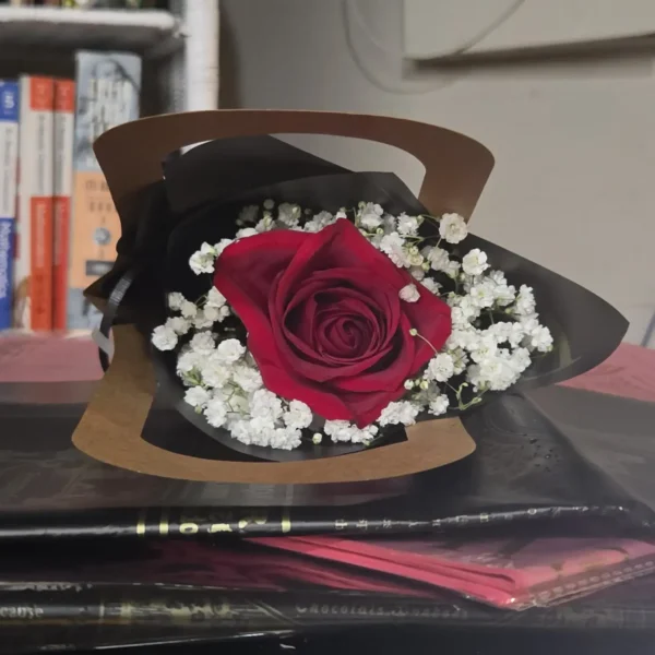 A single red rose wrapped in black and brown paper, surrounded by white baby’s breath, resting on a white surface with a softly blurred warm background.