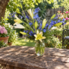 Pale yellow lilies, tall blue delphinium spires, and green fern foliage arranged in a clear glass ribbed vase.
