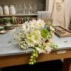White and cream bouquet with blush pink flowers, a green layered bloom, a dark-centered cream flower, delicate white filler flowers, and trailing green foliage.