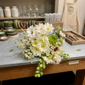 Anemone, Sweet Pea White Leopard Bouquet White and cream bouquet with blush pink flowers, a green layered bloom, a dark-centered cream flower, delicate white filler flowers, and trailing green foliage.