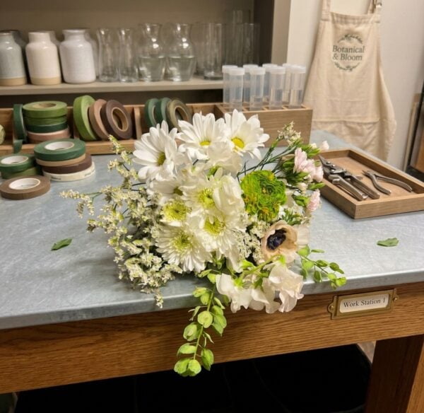 White and cream bouquet with blush pink flowers, a green layered bloom, a dark-centered cream flower, delicate white filler flowers, and trailing green foliage.