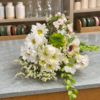 White and cream bouquet with blush pink flowers, a green layered bloom, a dark-centered cream flower, delicate white filler flowers, and trailing green foliage.