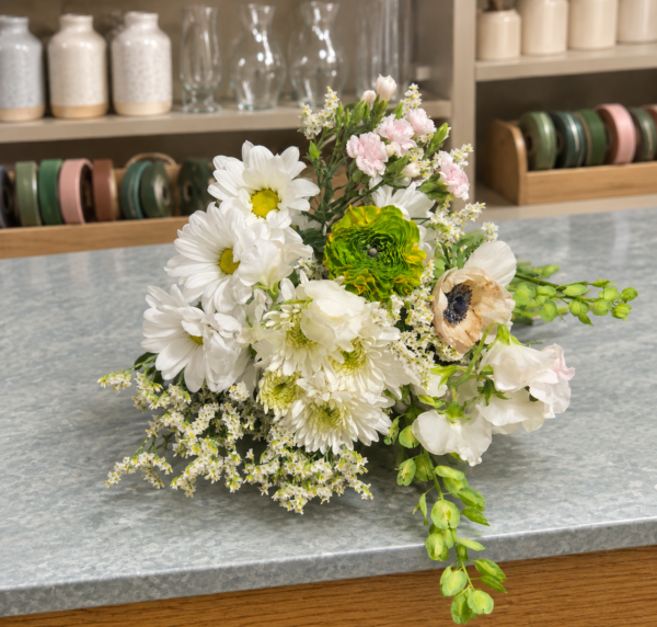 White and cream bouquet with blush pink flowers, a green layered bloom, a dark-centered cream flower, delicate white filler flowers, and trailing green foliage.